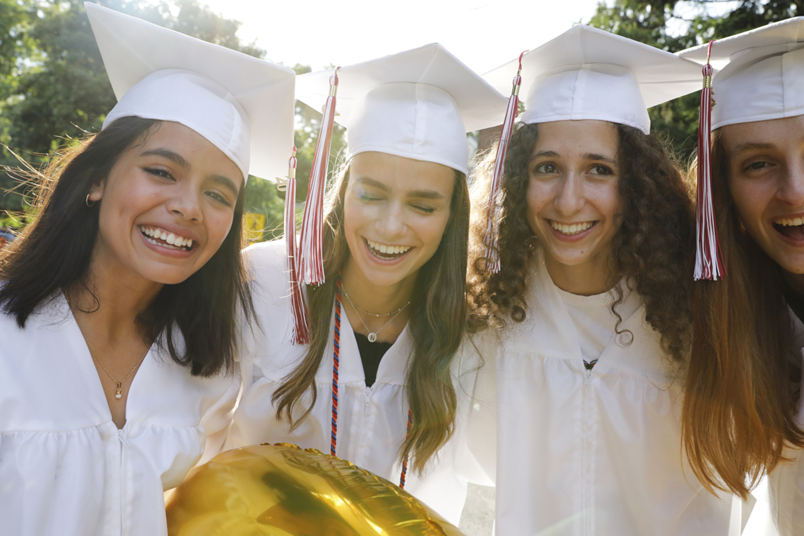 Group portrait of 4 teenage girls on their graduation day. They are all wearing white caps and gowns. They are standing outside on a street in their neighborhood. They are all smiling and having a wonderful time. There is a beautiful backlight on them. Waist up portrait.