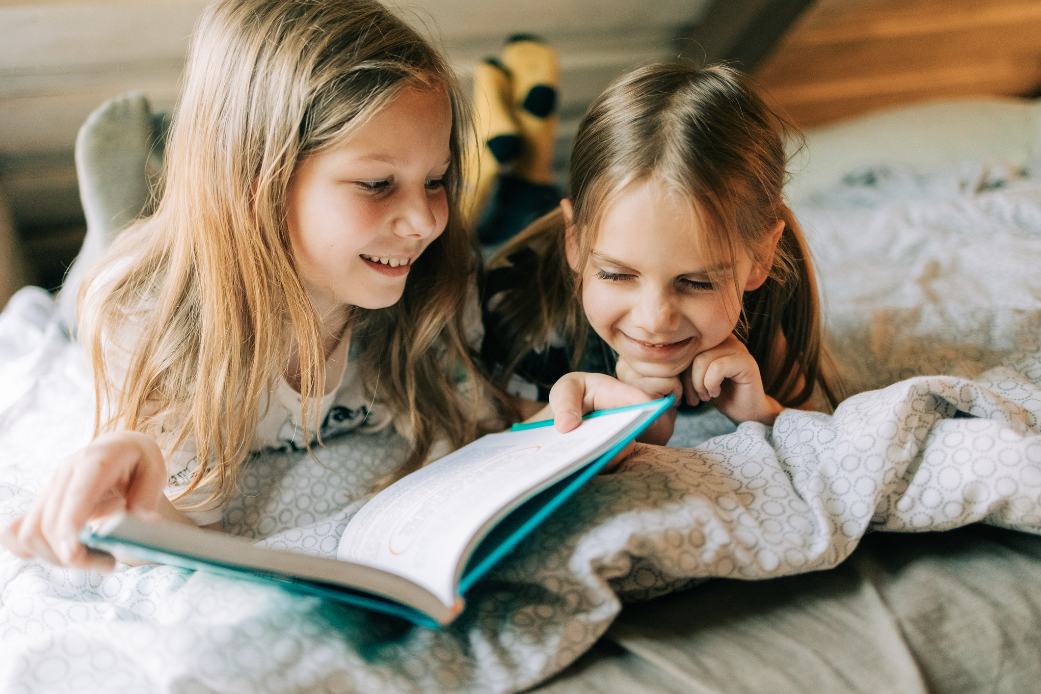 Two sisters are lying on bed reading book together