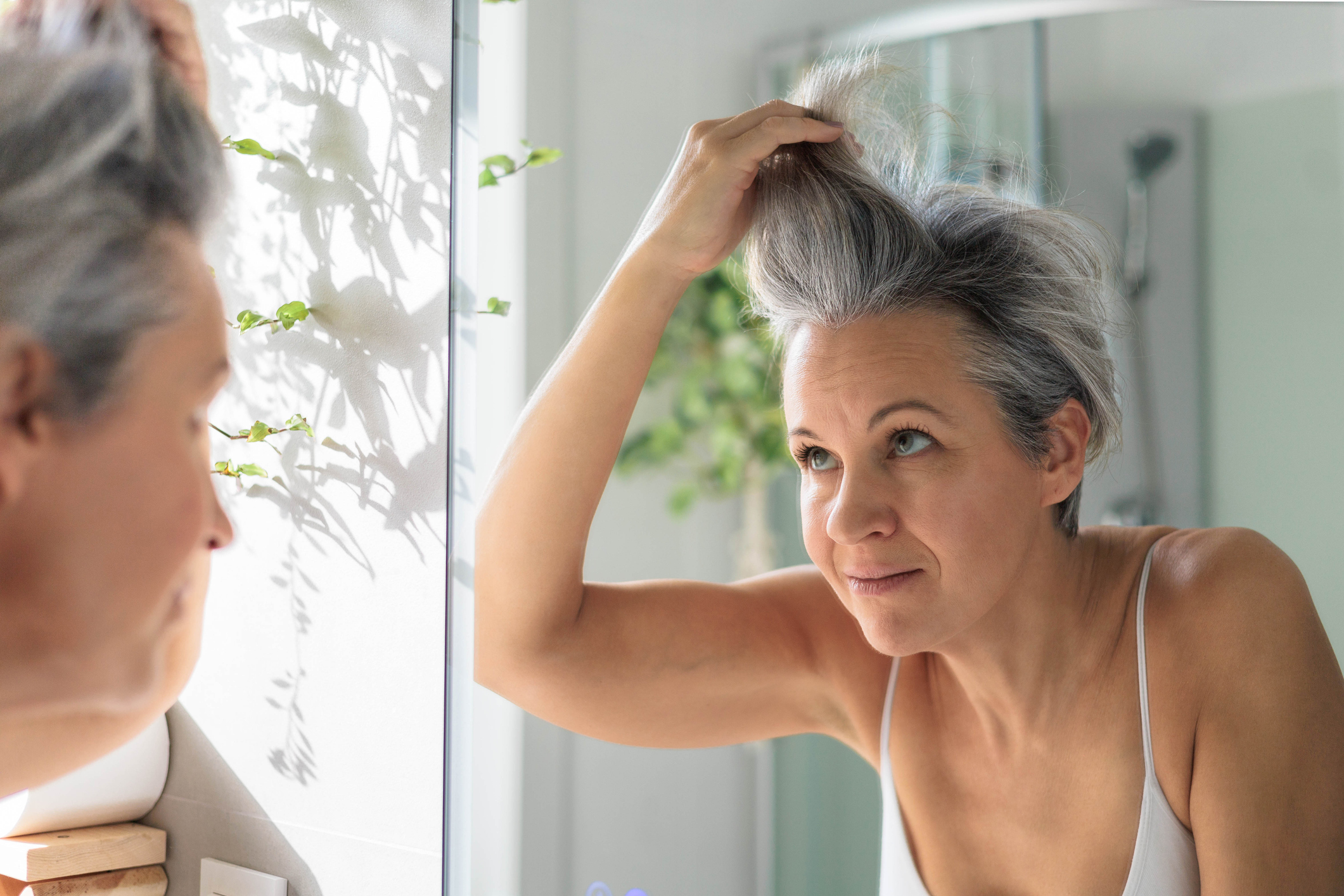 European beautiful and smiling middle-aged woman combing her gray hair and looking at her reflection in the mirror. Anti-age concept.